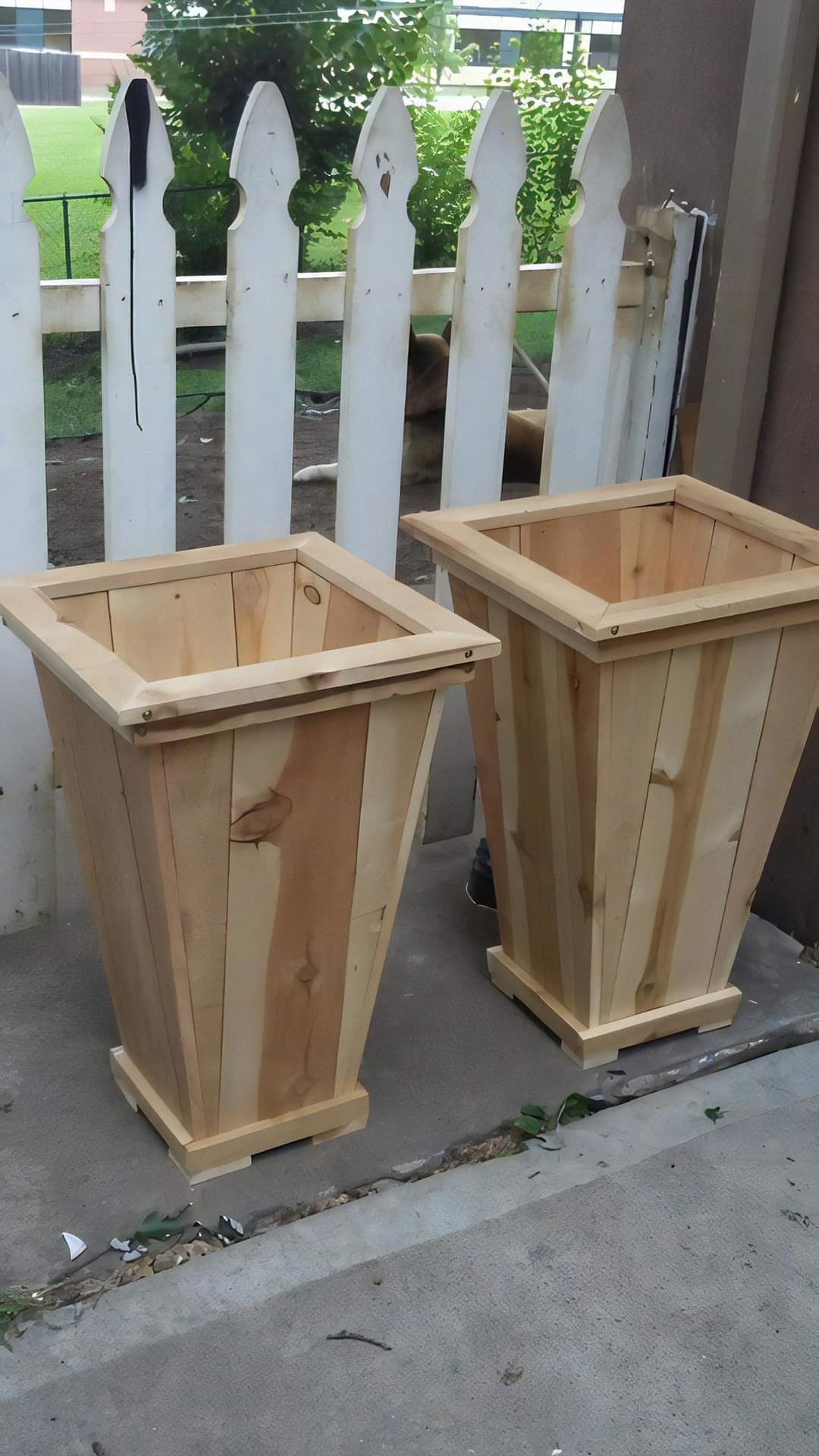 Two tapered wooden planter boxes standing beside a picket fence